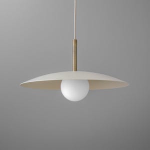 May include: A modern pendant light fixture with a white, saucer-shaped shade and a spherical white bulb. The fixture has a brass-colored stem and a white cord. The overall design is minimalist and elegant, suitable for various interior styles.