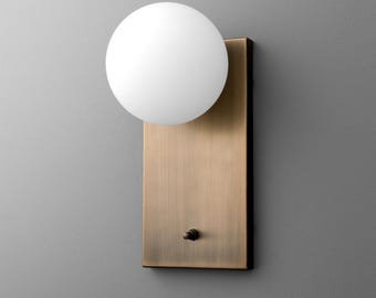 May include: A modern wall sconce featuring a white globe light fixture. The light is mounted on a rectangular, brushed brass-colored backplate. A small switch is located at the bottom of the backplate.