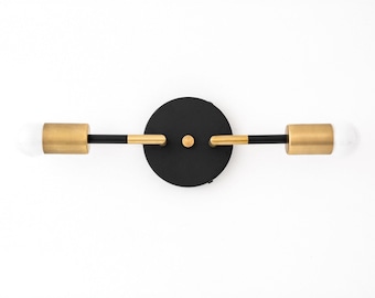 May include: Black and gold wall sconce with two light bulbs. The sconce has a round black base and two gold arms extending out with light bulbs at the ends.