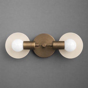 May include: A wall-mounted light fixture featuring a central circular antique brass base with two horizontal arms extending outward. Each arm holds a white spherical light bulb centered in front of a flat, circular, off-white metal backplate.