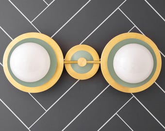 May include: A brass wall sconce with three circular lights. Each light has a white glass shade surrounded by a green ring and a gold ring.