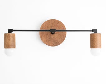 May include: A wall-mounted light fixture featuring a circular wooden backplate, a horizontal black metal bar, and two cylindrical wooden light sockets. Each socket holds a white globe light bulb, creating a balanced, linear design.