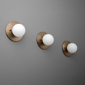 May include: Three wall sconces with a brass finish and a white globe light. The sconces are mounted on a gray wall. The light fixtures are round and modern.