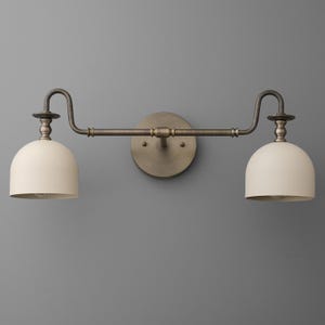 May include: A two-light wall sconce featuring a dark bronze-toned metal frame with a circular wall mount and two curved arms extending outward. Each arm supports a matte off-white dome-shaped shade directed downward, creating a balanced fixture.