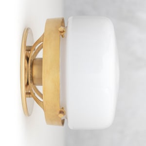 May include: A brass wall sconce with a white glass shade. The sconce has a circular base and a decorative design.