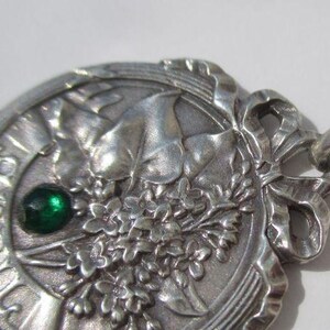 K&ouml;nnte beinhalten: Ein silberfarbener Metallanh&auml;nger mit kreisf&ouml;rmigem Design und einem Relief aus Blumenstrau&szlig; und Bl&auml;ttern. Das Wort APRIL ist am linken Rand eingepr&auml;gt. Ein kleiner, runder, dunkelgr&uuml;ner Edelstein ist in das Metall eingefasst. Oben befindet sich eine Schleife.