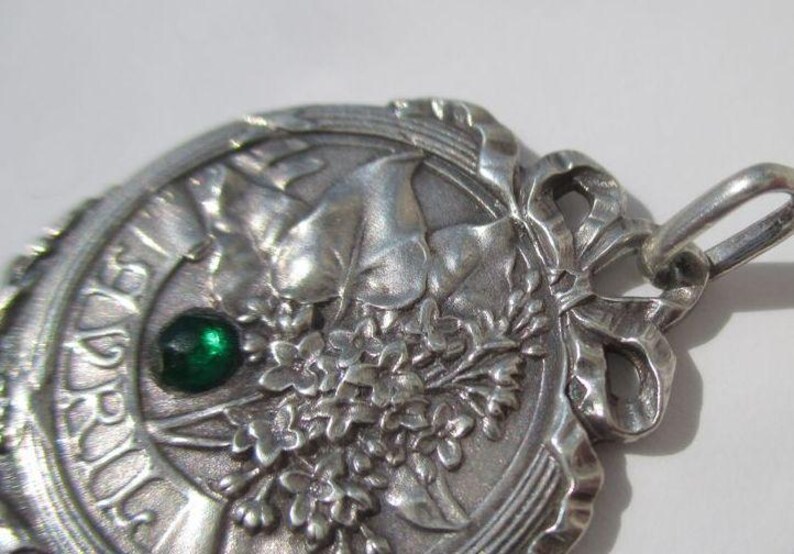 K&ouml;nnte beinhalten: Ein silberfarbener Metallanh&auml;nger mit kreisf&ouml;rmigem Design und einem Relief aus Blumenstrau&szlig; und Bl&auml;ttern. Das Wort APRIL ist am linken Rand eingepr&auml;gt. Ein kleiner, runder, dunkelgr&uuml;ner Edelstein ist in das Metall eingefasst. Oben befindet sich eine Schleife.