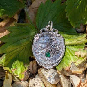 K&ouml;nnte beinhalten: Ein silberfarbener Metallanh&auml;nger mit gepr&auml;gtem Blumenmuster und einem kleinen, runden, leuchtend gr&uuml;nen Edelstein in der Mitte. Der Anh&auml;nger hat eine dekorative schleifenf&ouml;rmige &Ouml;se an der Oberseite und einen gewellten Rand mit Reliefdetails.