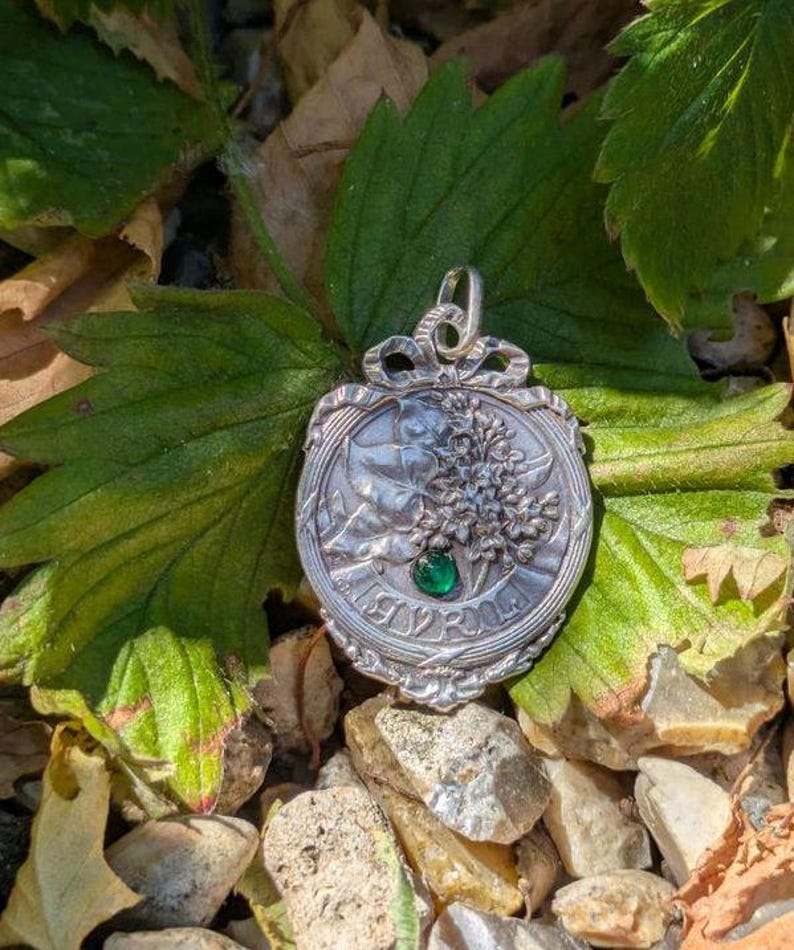 K&ouml;nnte beinhalten: Ein silberfarbener Metallanh&auml;nger mit gepr&auml;gtem Blumenmuster und einem kleinen, runden, leuchtend gr&uuml;nen Edelstein in der Mitte. Der Anh&auml;nger hat eine dekorative schleifenf&ouml;rmige &Ouml;se an der Oberseite und einen gewellten Rand mit Reliefdetails.