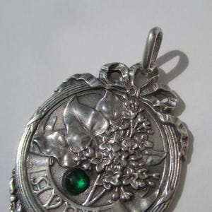 K&ouml;nnte beinhalten: Ein silberfarbener Metallanh&auml;nger mit einem Reliefdesign aus Bl&auml;ttern und Blumen sowie einem kleinen, runden, facettierten gr&uuml;nen Stein in der Mitte. Oben befindet sich eine schleifenartige Verzierung und am unteren Rand ist das Wort AVRIL eingepr&auml;gt.