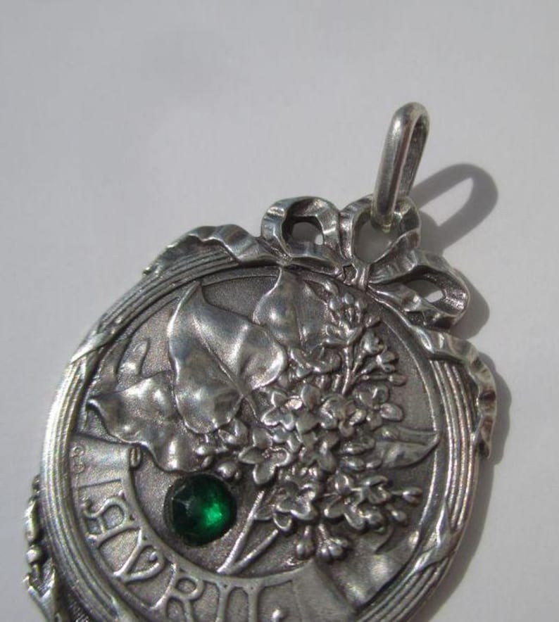 K&ouml;nnte beinhalten: Ein silberfarbener Metallanh&auml;nger mit einem Reliefdesign aus Bl&auml;ttern und Blumen sowie einem kleinen, runden, facettierten gr&uuml;nen Stein in der Mitte. Oben befindet sich eine schleifenartige Verzierung und am unteren Rand ist das Wort AVRIL eingepr&auml;gt.