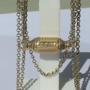 May include: A gold-colored necklace with a decorative cylindrical clasp. The necklace is displayed on a white stand. The clasp has intricate detailing and is connected to multiple strands of chain. The chains are made of small, rounded links.