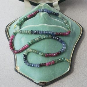 Op de afbeelding: Een veelkleurige edelstenen ketting met robijn, saffier en smaragd kralen. De ketting wordt getoond in een vintage, lichtgroene sieradendoos met een zwarte rand. De ketting is vastgezet met een gouden sluiting.
