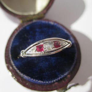 Peut inclure: Une bague argentée dotée d'un sertissage en forme de marquise avec une pierre claire ronde centrale, flanquée de deux pierres rouges carrées et de deux pierres claires plus petites sur les bords, le tout sur un anneau en métal détaillé.