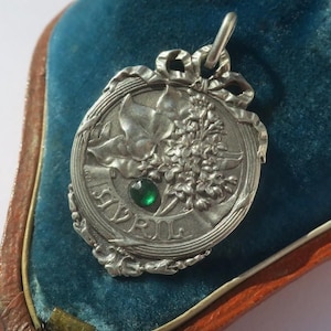 K&ouml;nnte beinhalten: Ein silberfarbener Metallanh&auml;nger mit einem Reliefdesign aus Bl&auml;ttern und Blumen sowie einer schleifenartigen Verzierung am oberen Rand. Ein kleiner, runder, facettierter gr&uuml;ner Stein ist in der Mitte eingelassen. Das Wort AVRIL steht auf einem Band.