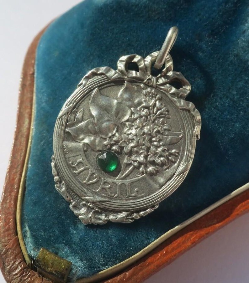 K&ouml;nnte beinhalten: Ein silberfarbener Metallanh&auml;nger mit einem Reliefdesign aus Bl&auml;ttern und Blumen sowie einer schleifenartigen Verzierung am oberen Rand. Ein kleiner, runder, facettierter gr&uuml;ner Stein ist in der Mitte eingelassen. Das Wort AVRIL steht auf einem Band.