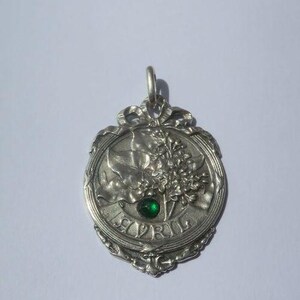 K&ouml;nnte beinhalten: Ein silberfarbener Metallanh&auml;nger mit einem Reliefdesign aus Blumenzweigen und Bl&auml;ttern. Das Wort AVRIL ist unten eingepr&auml;gt, und ein kleiner, runder, facettierter gr&uuml;ner Edelstein ist in die Mitte des Blumenarrangements eingesetzt. Der Anh&auml;nger hat einen dekorativen Rand und eine &Ouml;se zum Aufh&auml;ngen.
