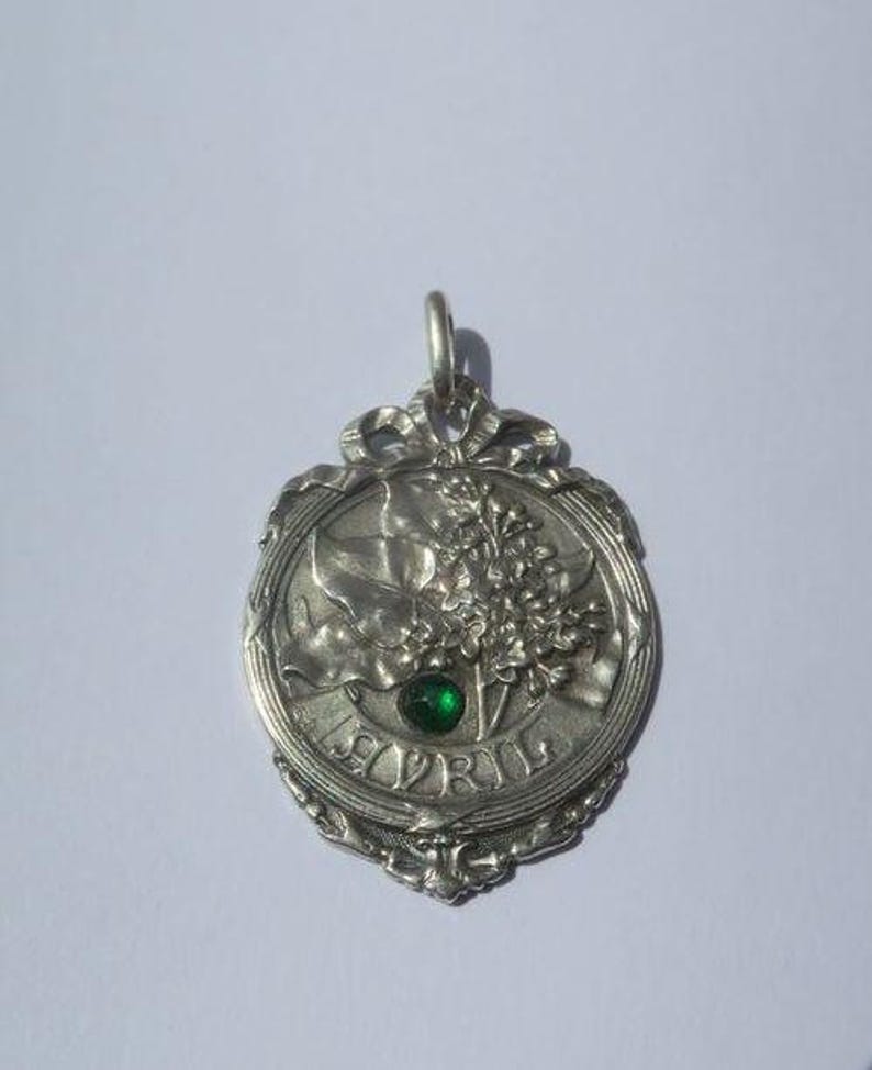 K&ouml;nnte beinhalten: Ein silberfarbener Metallanh&auml;nger mit einem Reliefdesign aus Blumenzweigen und Bl&auml;ttern. Das Wort AVRIL ist unten eingepr&auml;gt, und ein kleiner, runder, facettierter gr&uuml;ner Edelstein ist in die Mitte des Blumenarrangements eingesetzt. Der Anh&auml;nger hat einen dekorativen Rand und eine &Ouml;se zum Aufh&auml;ngen.
