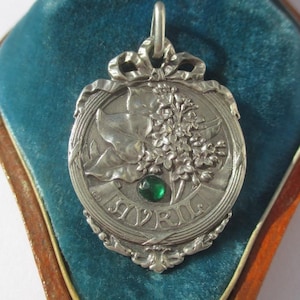 K&ouml;nnte beinhalten: Ein silberfarbener Metallanh&auml;nger mit einem Reliefdesign aus Bl&auml;ttern und Blumen sowie einem kleinen, runden, facettierten gr&uuml;nen Stein. Das Wort AVRIL ist unten im kreisf&ouml;rmigen Rahmen eingepr&auml;gt, der von einer schleifenf&ouml;rmigen &Ouml;se gekr&ouml;nt wird.