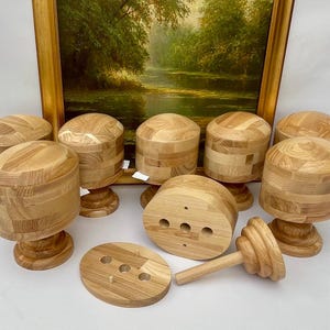 May include: A selection of unfinished wooden hat blocks and components. The blocks are cylindrical with a rounded top, and some are mounted on pedestals. The wood has a natural, light brown colour. A painting in a gold frame is in the background.