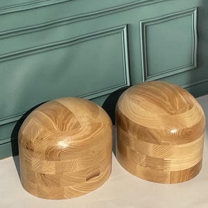 Könnte beinhalten: Zwei kuppelförmige Hutformen aus hellem Holz mit sichtbarer Maserung. Jede Form verfügt über eine glatte, abgerundete Oberseite und eine zylindrische Basis, die aus mehreren zusammengefügten Holzsegmenten mit natürlichem Finish besteht.