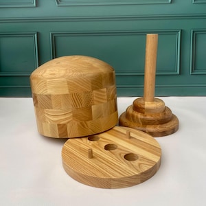 Puede incluir: Un juego de bloques de madera para sombreros, que incluye un bloque en forma de cúpula, una base redonda con clavijas y un soporte. El juego está hecho de madera clara con un acabado natural, mostrando la veta de la madera.