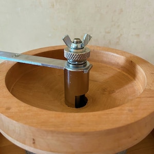 May include: A wooden grain mill with a metal grinder and a silver knob. The grinder is attached to a wooden base with a gray stone base.