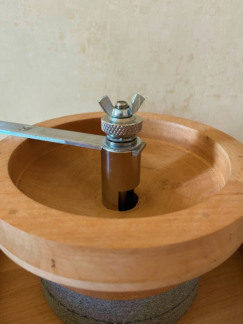 May include: A wooden grain mill with a metal grinder and a silver knob. The grinder is attached to a wooden base with a gray stone base.