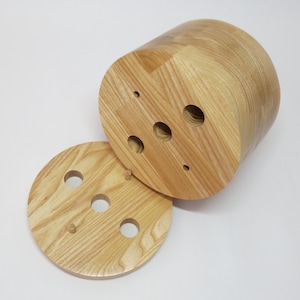 Puede incluir: Un contenedor de almacenamiento de madera con tapa. El contenedor tiene forma cilíndrica y está hecho de madera de color claro. La tapa tiene varios agujeros redondos cortados.