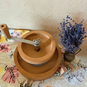May include: A wooden hand-crank grain mill with a gray stone grinding wheel. The mill is made of two stacked wooden rings with a crank handle on the top ring. The mill is sitting on a table with a floral pattern.