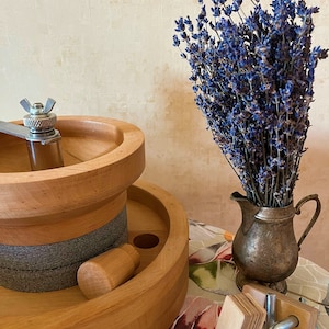 May include: A wooden grinding wheel with a gray stone and a wooden handle. A bouquet of dried lavender flowers is in a silver pitcher on a table with a floral pattern.