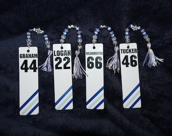 May include: Four white rectangular bookmarks with names and numbers, including "GRAHAM 44", "LOGAN 22", "DILAURENTIS 66", and "TUCKER 46". Each has a blue and silver beaded loop and tassel.