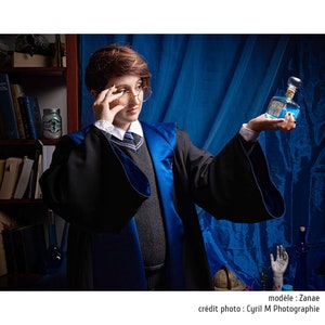 May include: A person wearing a black and blue robe with a gray sweater, white collared shirt, and a striped tie. They are holding a clear bottle with blue liquid and a decorative stopper. The background includes a bookshelf and blue fabric.