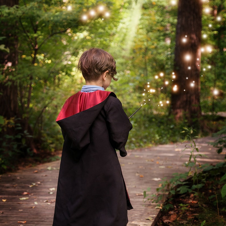 May include: A child wearing a black robe with a red hood and holding a wand, standing on a wooden path in a forest. The scene is illuminated by soft, glowing lights, creating a magical atmosphere. The child is facing away from the camera.