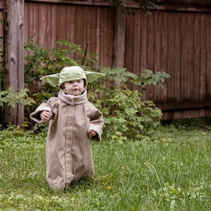 Puede incluir: Un ni&ntilde;o peque&ntilde;o vestido con una t&uacute;nica marr&oacute;n y un sombrero verde de Yoda camina por un campo de hierba.