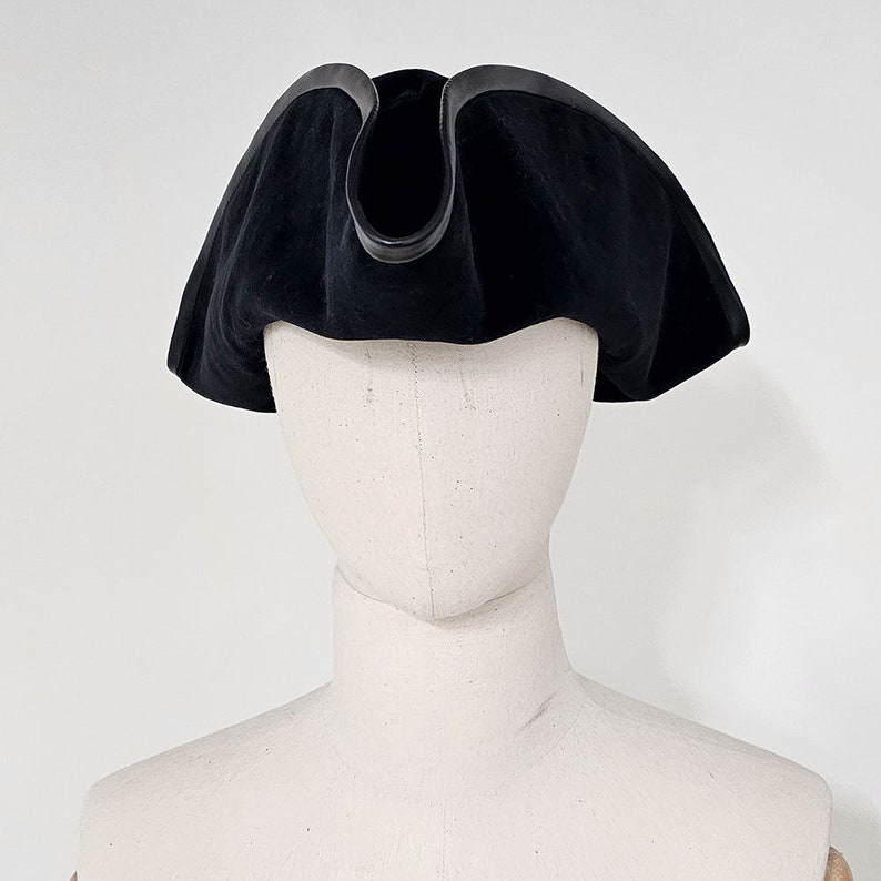 May include: A black velvet tricorn hat with a black leather trim. The hat is on a white mannequin head.