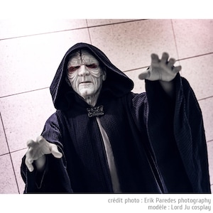 May include: A person dressed as Darth Sidious from Star Wars, wearing a black hooded robe with a white undergarment. The character has a pale face with red eyes and a dark expression.