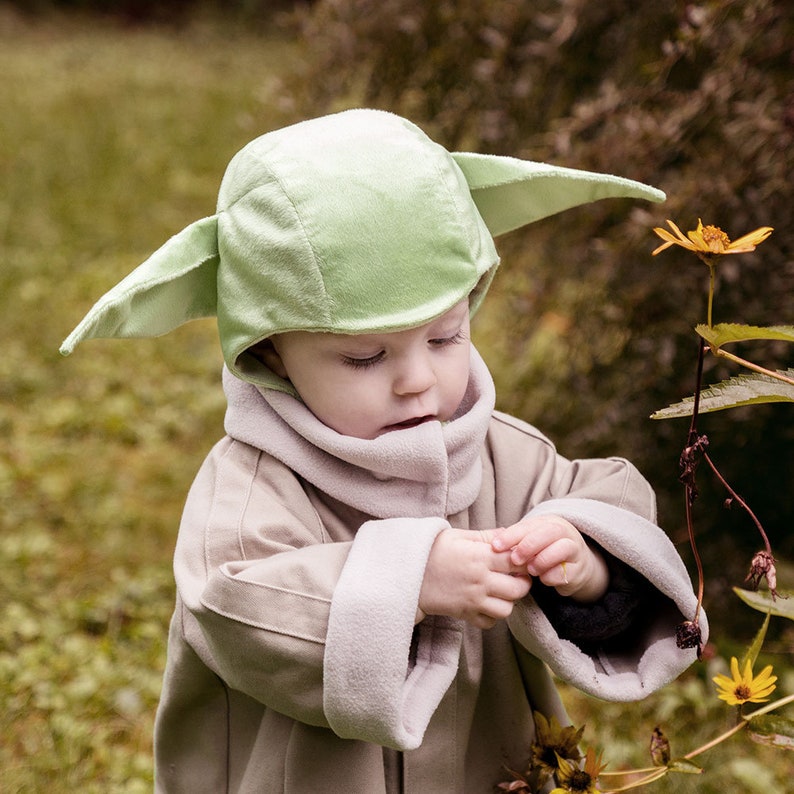 Puede incluir: Un ni&ntilde;o peque&ntilde;o con un disfraz verde de Yoda, mirando hacia abajo a una flor. El disfraz incluye un sombrero con orejas grandes y una t&uacute;nica marr&oacute;n.
