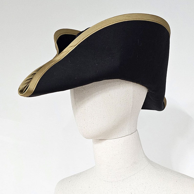 May include: A black tricorn hat with a gold trim. The hat is angled to the side and is sitting on a white mannequin head.