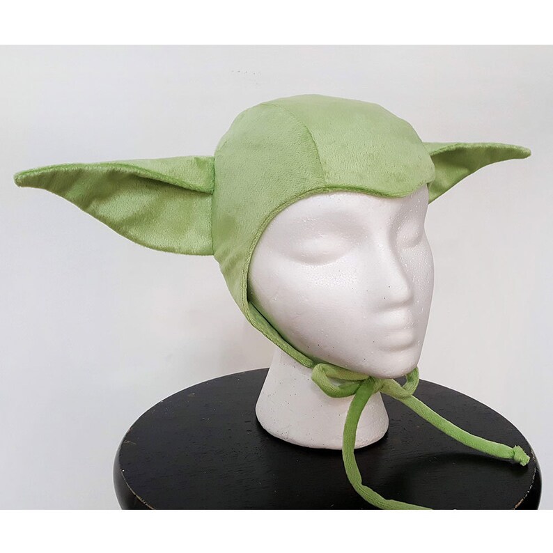 May include: A green plush hat with large ears, resembling a popular fictional character. The hat has a tie at the back for a secure fit.
