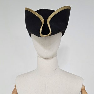 May include: A black tricorn hat with a gold trim. The hat is on a white mannequin head.