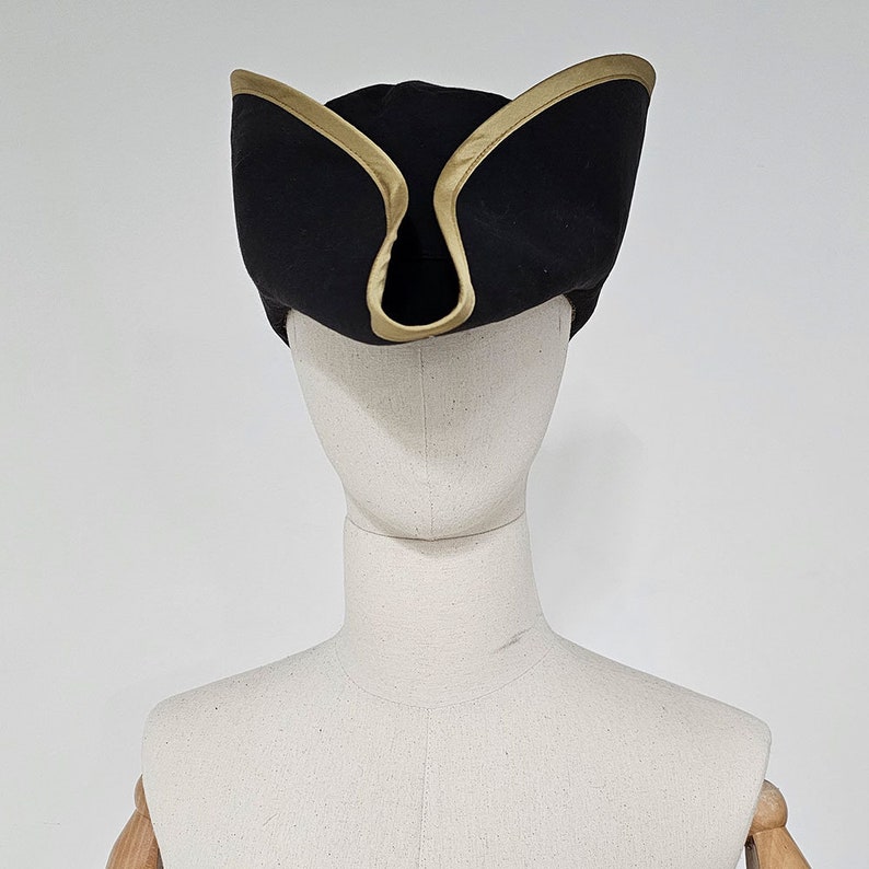 May include: A black tricorn hat with a gold trim. The hat is on a white mannequin head.