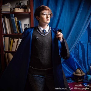 May include: A person wearing a dark gray sweater, white collared shirt, and blue and black robe. The robe has a blue satin lining. The person is wearing glasses and a tie with blue stripes. A silver pin is attached to the sweater. A bookshelf and blue curtain are in the background.