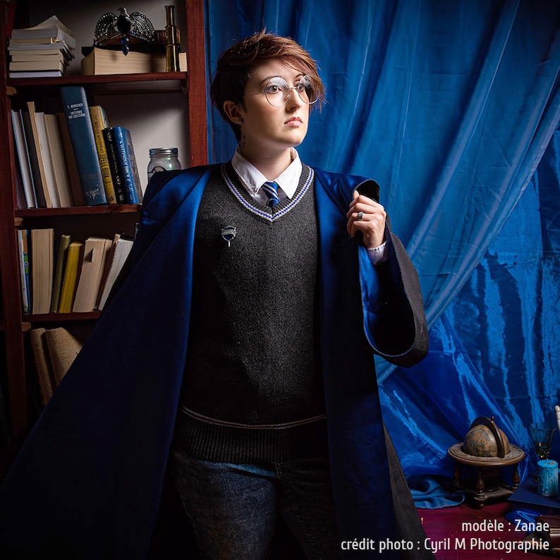 May include: A person wearing a dark gray sweater, white collared shirt, and blue and black robe. The robe has a blue satin lining. The person is wearing glasses and a tie with blue stripes. A silver pin is attached to the sweater. A bookshelf and blue curtain are in the background.