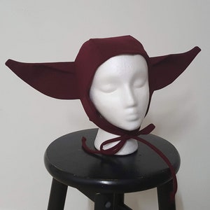 May include: A burgundy hooded hat with large pointed ears. The hat is made of a soft, stretchy fabric and has a simple design.