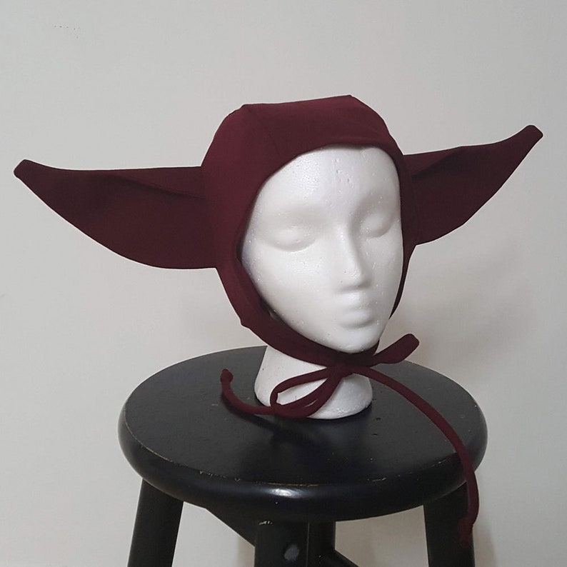 May include: A burgundy hooded hat with large pointed ears. The hat is made of a soft, stretchy fabric and has a simple design.
