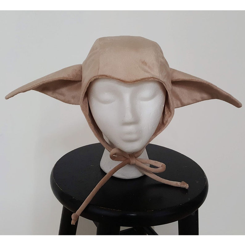 May include: A beige felt elf hat with large pointed ears and a tie closure. The hat is worn on a white mannequin head.