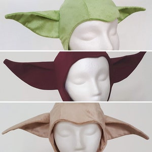 May include: Three different coloured Yoda-style hats. The hats are made of soft fabric and have large, floppy ears. The hats are green, maroon, and beige.