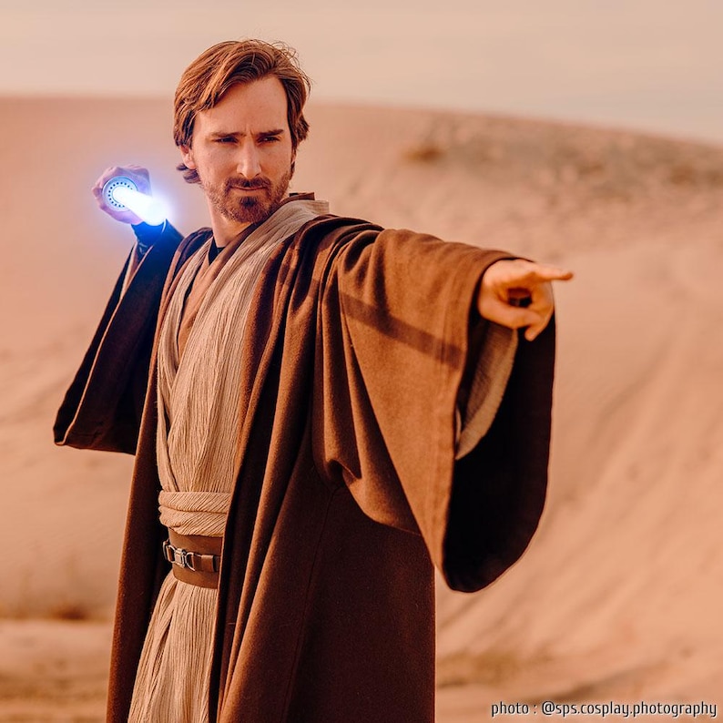 May include: A person dressed as a Jedi Knight, wearing a brown robe and holding a blue lightsaber, stands in a desert landscape. The person is pointing their right hand forward.