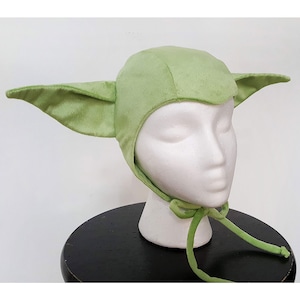 Puede incluir: Sombrero de Yoda de felpa verde con orejas grandes y una corbata en la parte posterior. El sombrero est&aacute; en una cabeza de maniqu&iacute; blanca.